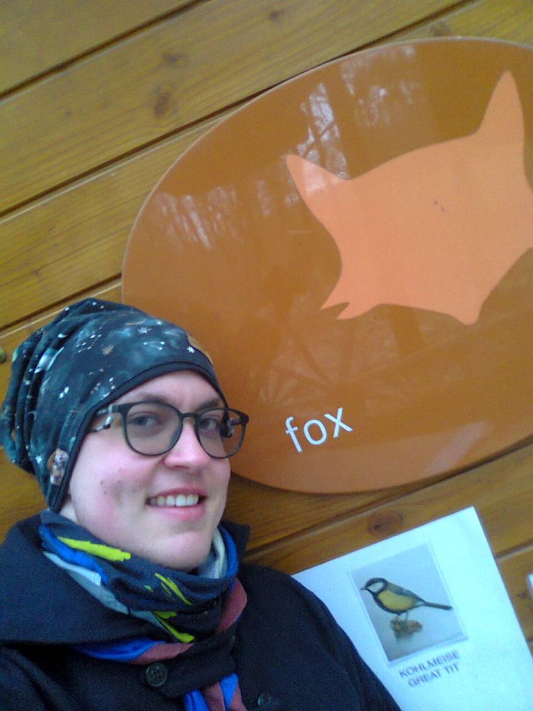 Ein junger Mann steht mit Juscha und Pfadfinderhalstuch vor einem Schild mit der Aufschrift "Fox"