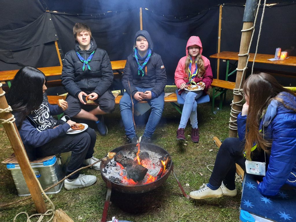 Fünf Pfadfinder*innen sitzen in einem Zelt um ein Lagerfeuer herum