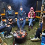 Fünf Pfadfinder*innen sitzen in einem Zelt um ein Lagerfeuer herum