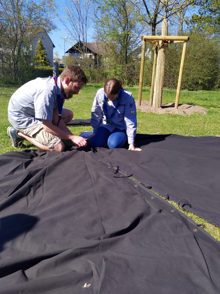 Ein Pfadfinder und eine Jungspfadfinderin knien auf der Wiese und knöpfen schwarze Zeltplanen aneinander