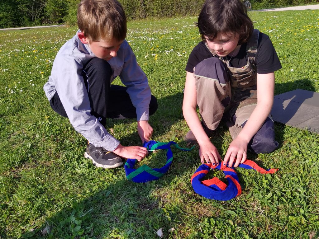 Ein Junge und ein Mädchen knieen auf einer Wiese und knoten Pfadfinder*innen Halstücher