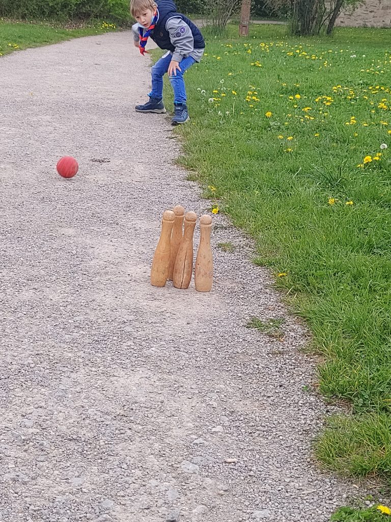 Ein Junge steht auf einem Schotterweg. Vor ihm rollt eine rote Kugel auf vier hölzerne Kegel zu.