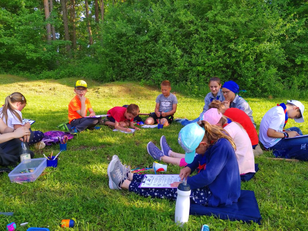 Kinder sitzen im Kreis in der schattigen Wiese. Sie malen auf Klemmbrettern.