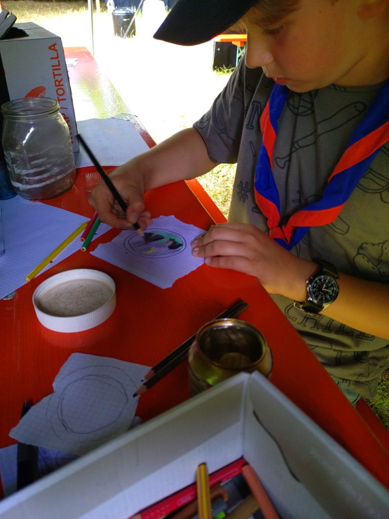 Ein Pfadfinder sitzt malend an einem Biertisch. Um ihn rum liegen Stifte und Papierschnipsel.