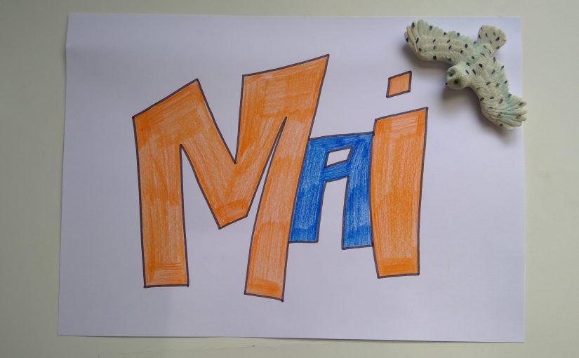 Ein Blatt mit dem Wort "Mai" in orangnen und blauen Buchstaben liegt auf einem weißen Schreibtisch. Daneben liegt eine Schneeeulen