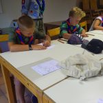 Zwei Jungen und ein Mädchen sitzen an einem weißen Tisch und malen/schreiben etwas auf Papier.