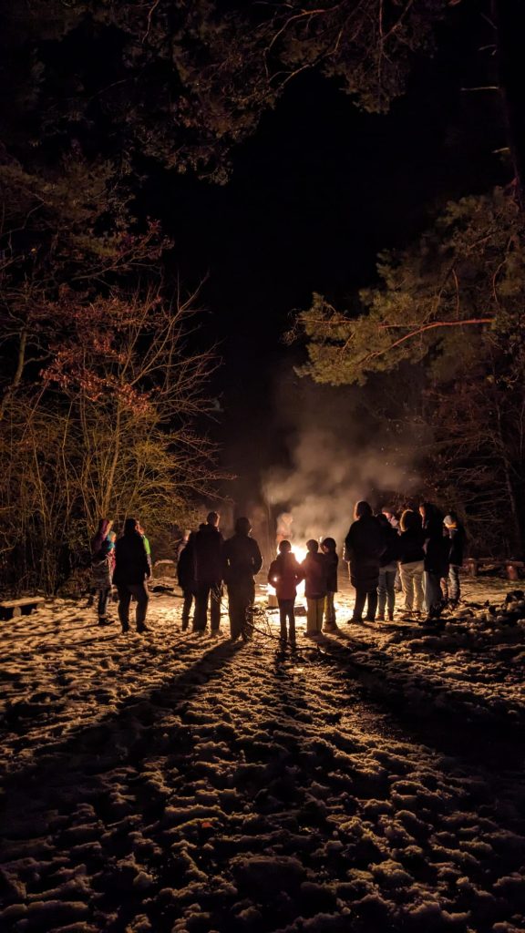 Kinder verschiedenen Alters stehen im Kreis um ein Feuer herum, dass in der Dunkelheit die einzige Lichtquelle darstellt